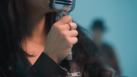 primer plano de un cantante sin rostro con una chaqueta de cuero, sosteniendo un micrófono vintage con una mano adornada con anillos. en el fondo azul borroso, un hombre es visible