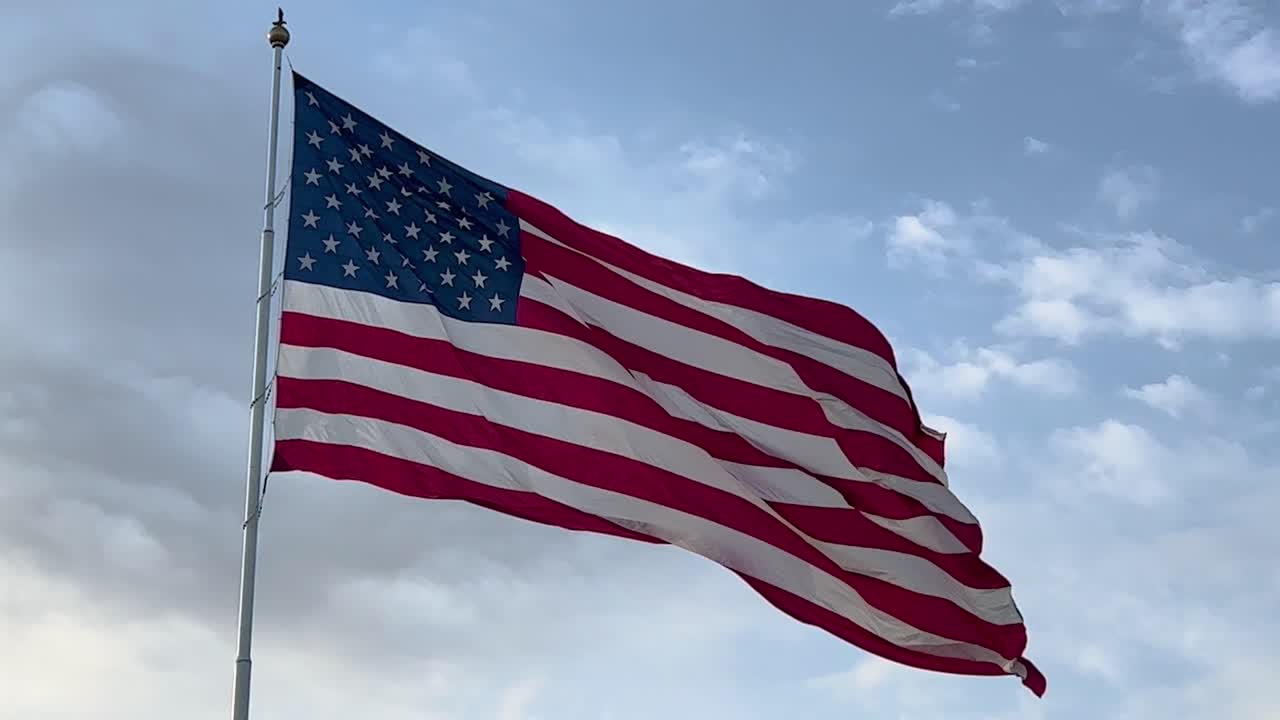 gran bandera estadounidense en cámara lenta soplando en el viento contra un cielo azul soleado y nubes blancas a finales de la tarde