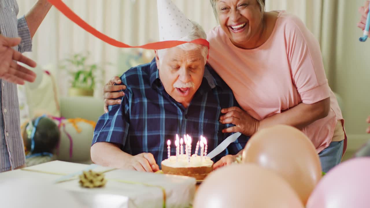 Uomo anziano caucasico e felice a una festa di compleanno con torta, insieme ad amici anziani di diverse etnie, al rallentatore.