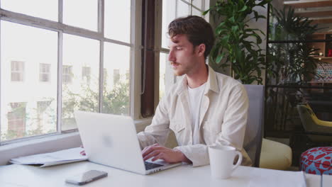 Millennial-white-male-creative-sitting-at-a-desk-using-laptop-in-a-casual-office,-panning-shot
