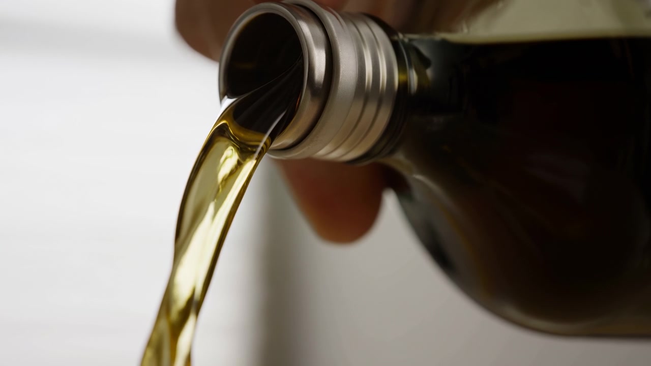 Una bottiglia di vetro scuro si inclina in un primissimo piano, rilasciando un liquido dorato traslucido attraverso il tappo metallico in un flusso laminare liscio, con riflessi lucidi, luci calde e un aspetto da studio pulito