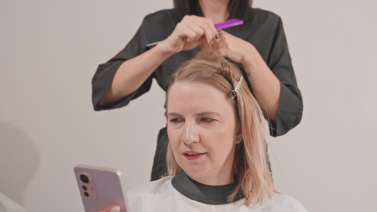 Proses penataan rambut *highlight* di salon oleh wanita kulit putih dengan penata rambut yang membagi rambut, menempelkan *foil*, menyisir rambut, menjepit bagian rambut saat klien menggulir *smartphone* dengan latar belakang studio netral, serta menggunakan pelindung