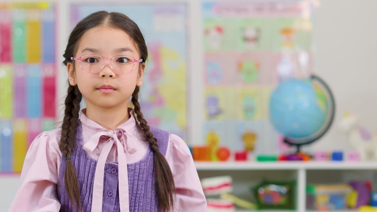 Smiling Asian girl holds green foam six in bright classroom with colorful posters and globe