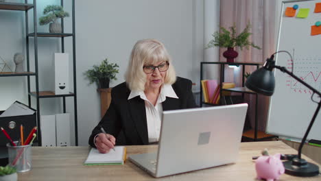 Senior-mature-older-business-office-woman-elearning-online-writing-notes-from-laptop,-live-webinar