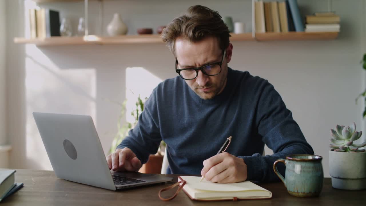 Mann i genser og briller taster, tar frem notatbok på hjemmekontoret, skriver notater for jobb med laptop og penn