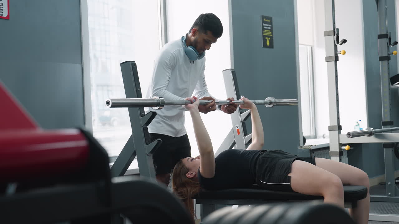 woman lifting barbell on bench while male trainer supports during indoor gym workout session, with modern fitness equipment visible in foreground and sunlight entering through large windows