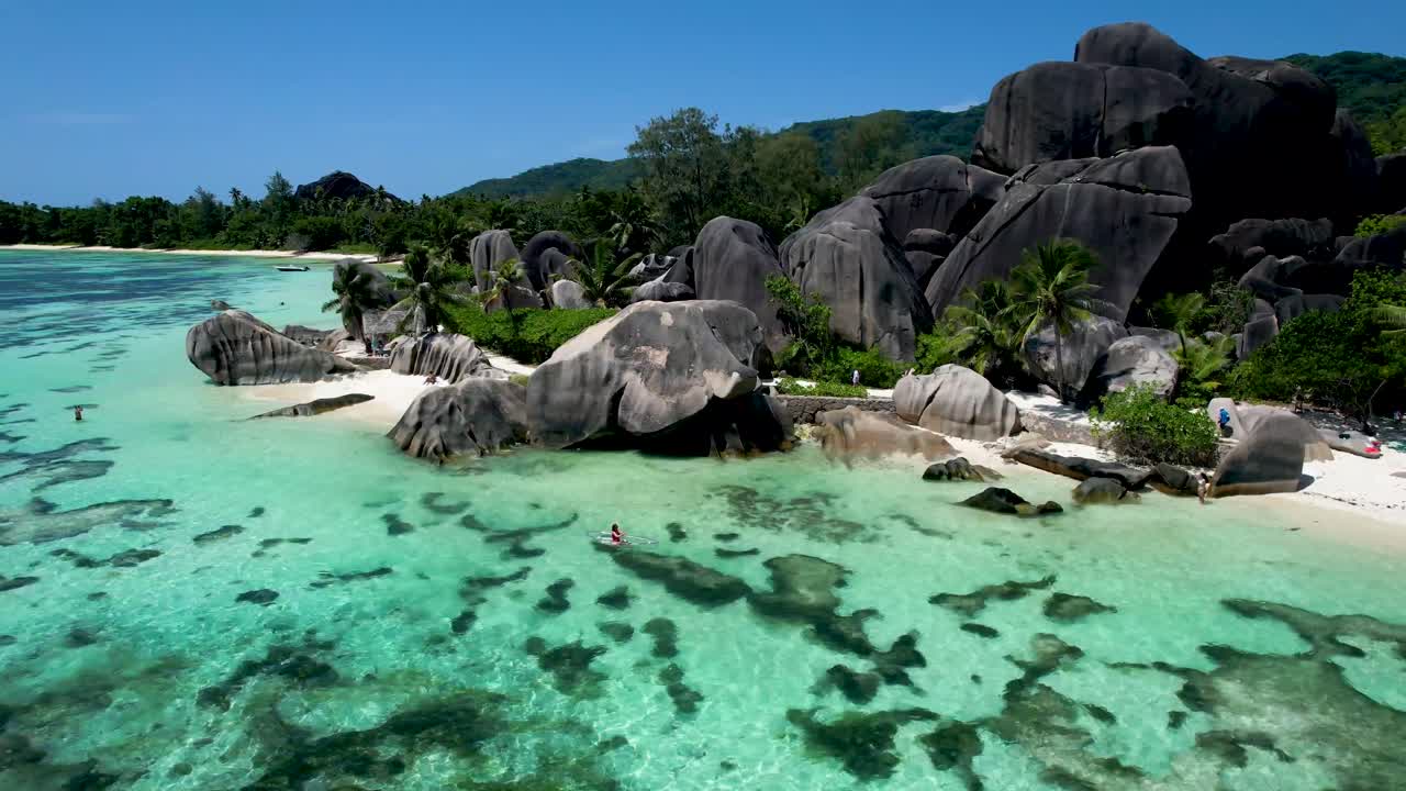 바위 야자나무 하얀 모래가 있는 상징적인 세이셸 해변의 공중 패닝 뷰 투명한 바닥 카약을 탄 여성