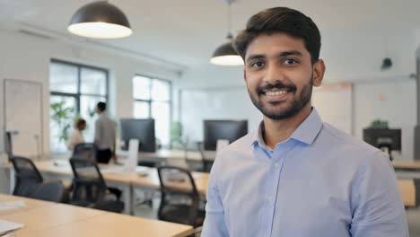 young confident indian entrepreneur smiling in modern startup company office with desks, computers and big windows, he is wearing a blue shirt and he has beard and dark hair