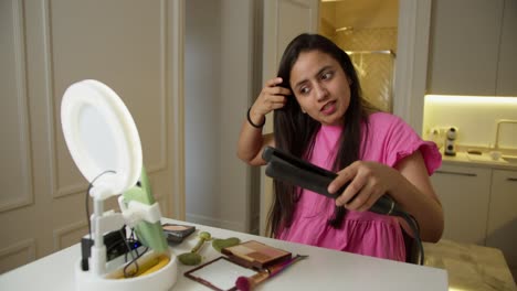 una feliz chica india con un vestido rosa dirige su blog de belleza y explica en un video cómo enderezar su cabello correctamente usando un hierro de enderezar especial en un apartamento moderno en línea