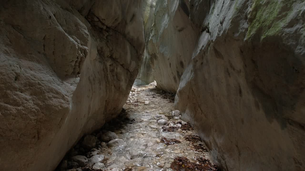 A stream flows through a narrow rock canyon The walls are tall and close together with light coming from above The water is clear flowing over rocks
