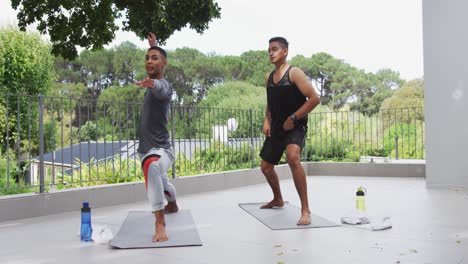 Mixed-race-gay-male-couple-standing-on-terrace-practicing-yoga