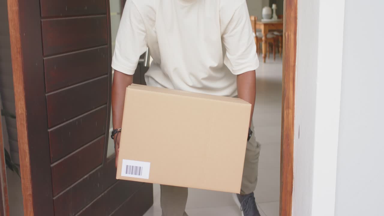 African American man retrieving cardboard box at doorstep, bending, lifting, carrying inside