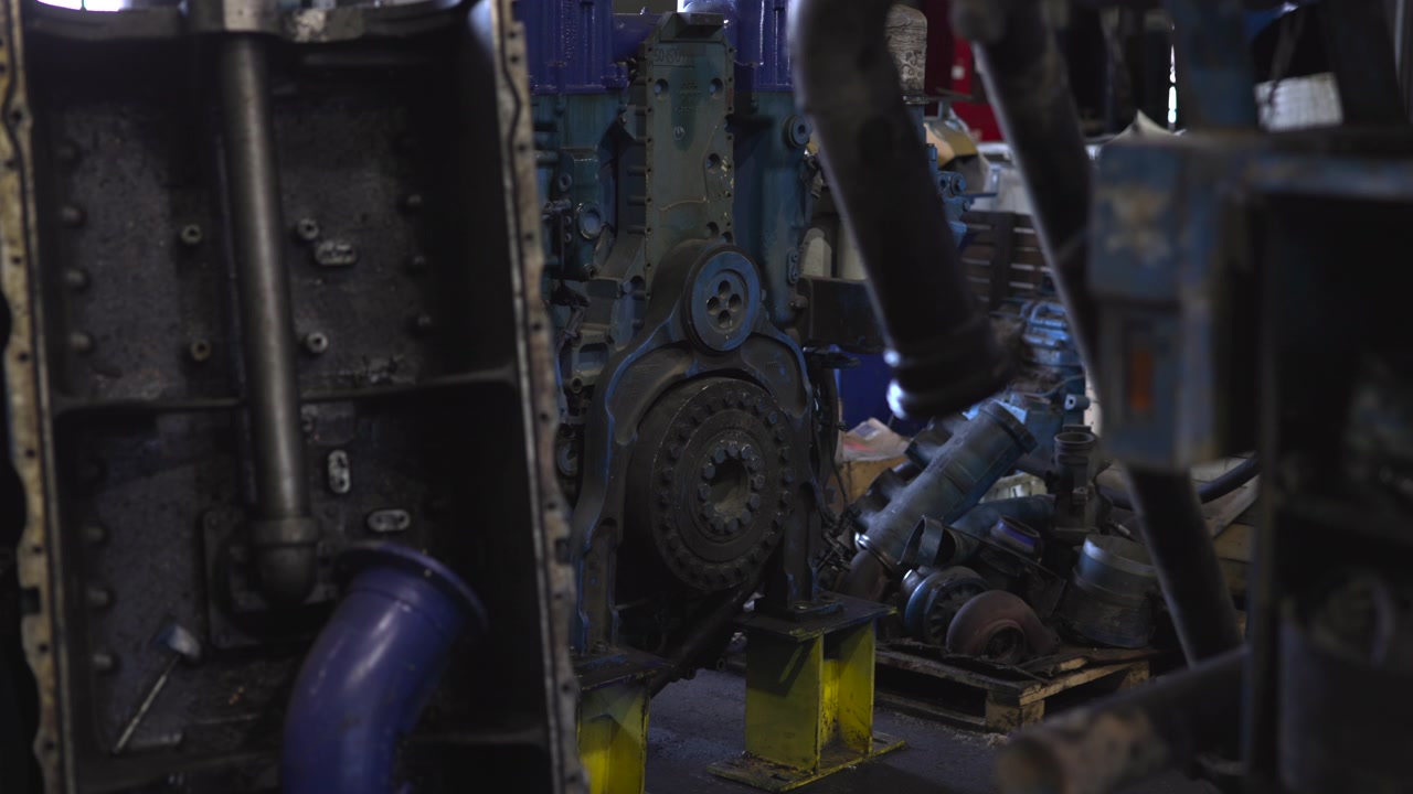 Disassembled blue-painted engine housing with exposed gear and pulley mounted on bright yellow metal supports inside cluttered industrial workshop, showing worn surfaces, tools, grime and repair