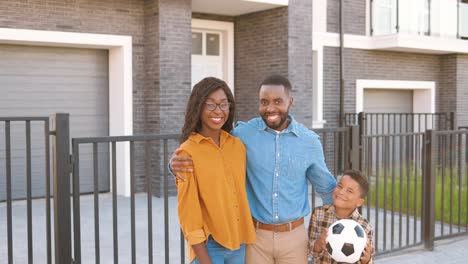 Retrato-De-Una-Familia-Afroamericana-Feliz-Con-Un-Niño-Lindo-Parado-En-La-Calle-Cerca-De-La-Casa-En-El-Suburbio-Y-Sonriendo-A-La-Cámara