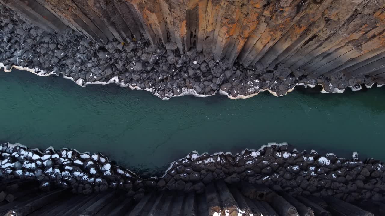 A unique aerial top-down perspective captures the stunning geological formations of Studlagil Canyon, revealing a turquoise river carving its way between towering, hexagonal basalt columns