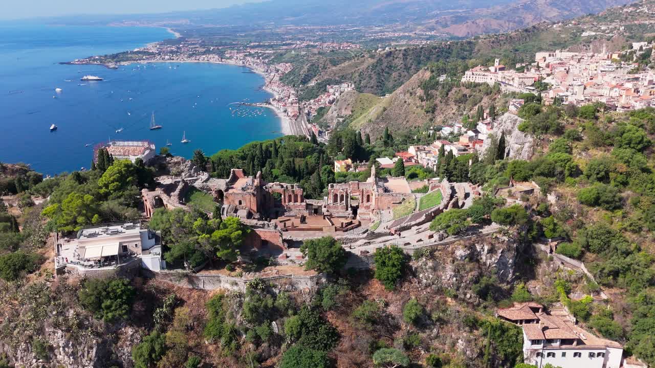Drone orbit shot from right to left around Taormina Amphitheatre, showing the historic structure with town rooftops and coastline in the background