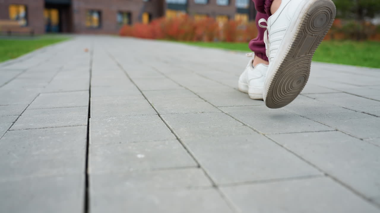 Leg view of trainer wearing white sneakers and burgundy sports suit walking along gray tiled pathway outside fitness center capturing dynamic stride and urban workout movement