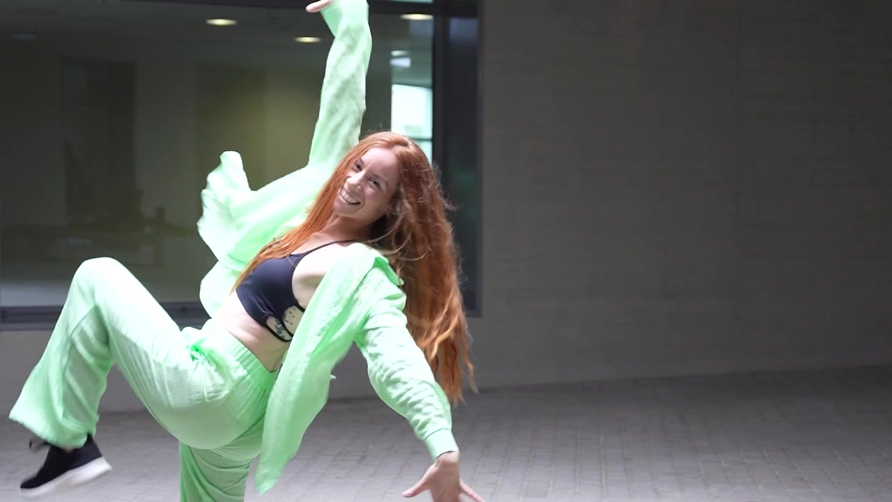 A woman is energetically dancing in a vibrant green outfit in a lively urban environment