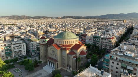 uhd 4k drone shot of church of saint panteleimon of acharnai in athens, greece. greek orthodox cathedral with green dome and red roof, surrounded by dense urban cityscape at golden hour.