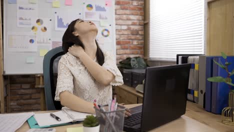 portrait of a stressed asian female employee is exercising her stiff neck muscles while working on the laptop at desk in a bright loft office.