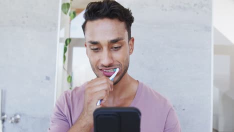 Hombre-Birracial-Feliz-Cepillándose-Los-Dientes-En-El-Baño-Por-La-Mañana-Y-Usando-Un-Teléfono-Inteligente,-Cámara-Lenta