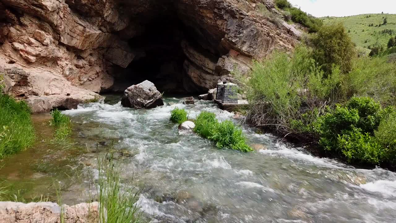 A drone shot over flowing water nearing the entrance of a cavern. (slow motion)