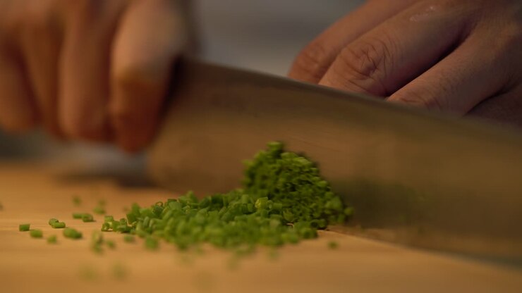 Mani di chef professionista che affettano la parte verde del cipollotto a piccole fette usando un coltello da cucina e un tagliere di legno, erba cipollina, cipollotti o cipolle verdi come ingrediente vegetale, primo piano estremo 4K.