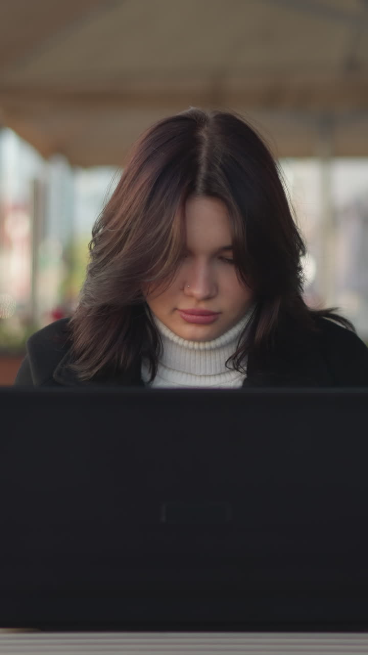 freelancer sentado al aire libre trabajando en una computadora portátil con expresión concentrada, ambientado en un entorno urbano con luz natural, muebles modernos y fondo borroso de edificios y flores