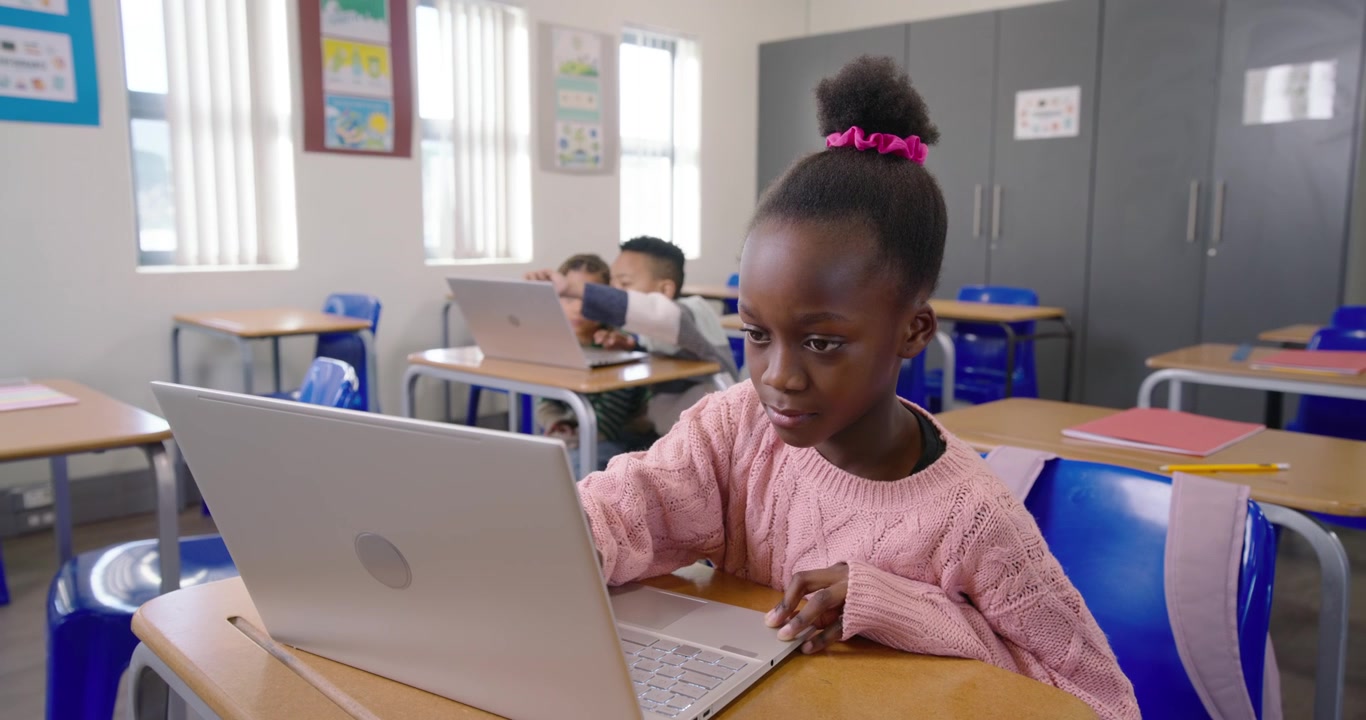 Bambina con maglione rosa che fa i compiti su un laptop bianco, la telecamera si allontana rivelando i compagni