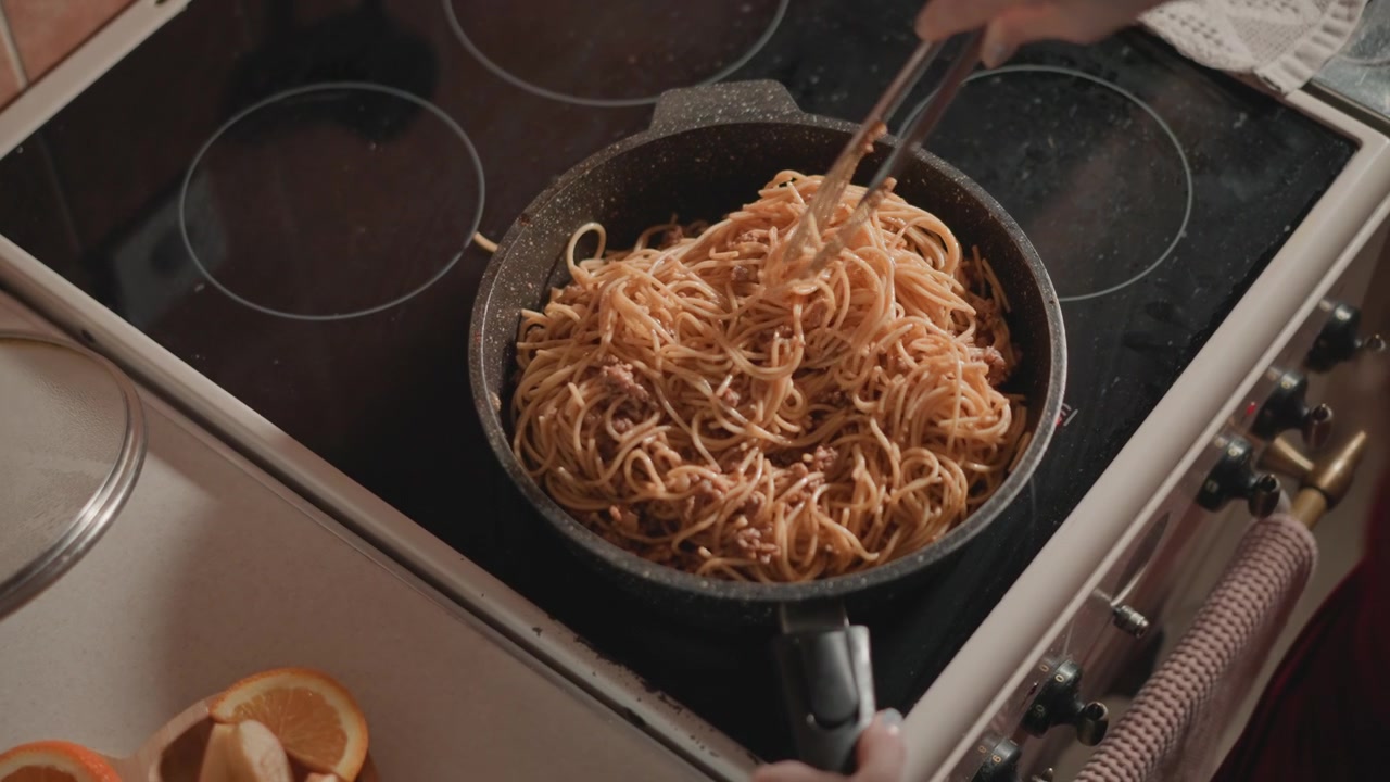 Primo piano dall'alto di noodles cotti mescolati e sollevati in una padella antiaderente scura usando una spatola di metallo, movimento mostrato da fili di noodles sollevati e utensile angolato mentre mano e tessuto rosso