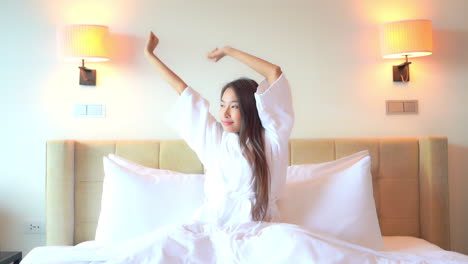 아침에 일어나기 위해 머리 위로 팔을 뻗어 흰 가운을 입고 침대에 앉아 있는 젊은 매력적인 아시아 여성