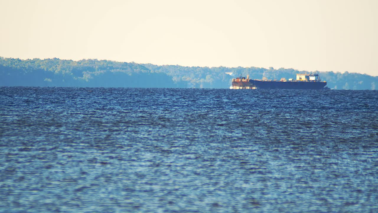 Telephoto clip of a cargo freighter gliding across a calm bay at sunset with forested shoreline in the background. Peaceful maritime and transport scene.