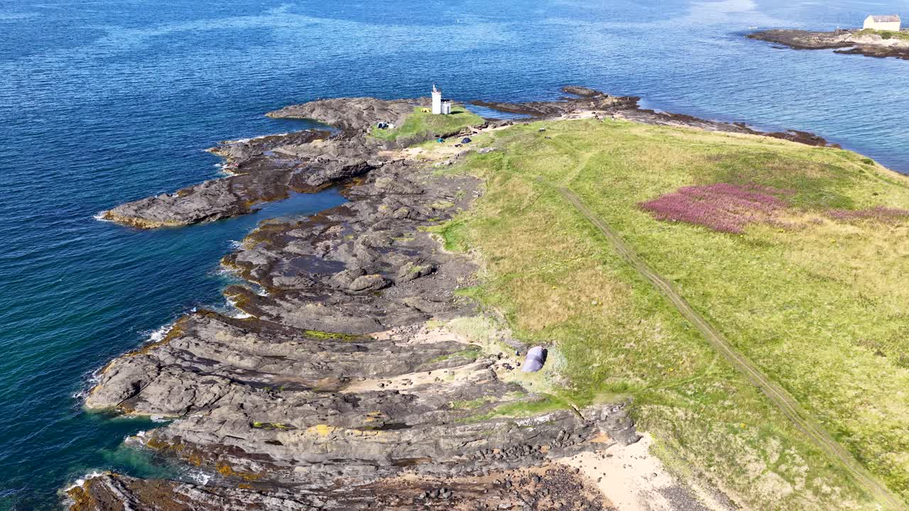 Drone glides over rocky shoreline toward lighthouse, bright daylight, vibrant green grass, blue sea
