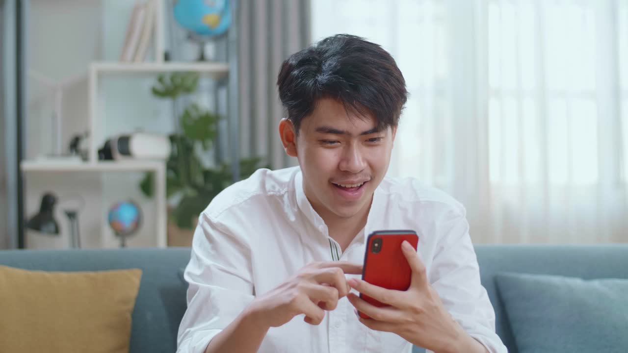 Happy Asian Man Celebrating While Using Smartphone In The Living Room