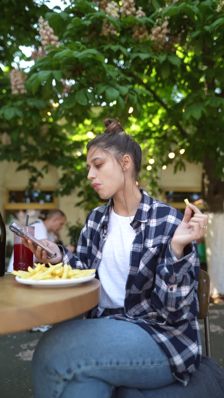 mujer joven comiendo papas fritas y usando su teléfono en un café al aire libre