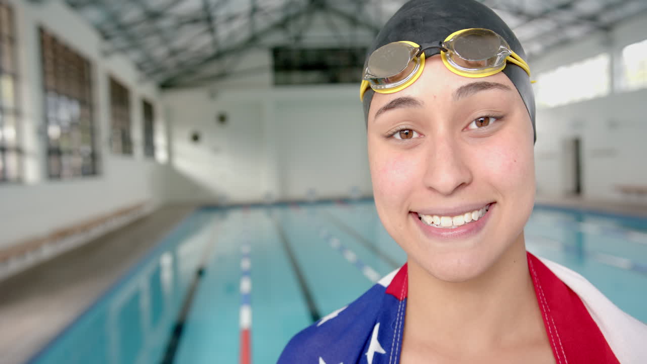 Smiling female swimmer with goggles and flag standing by indoor swimming pool, copy space