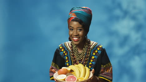 joven afroamericana alegre con turbante y ropa tradicional sonriendo alegremente a la cámara mientras baila y sostiene un plato con frutas