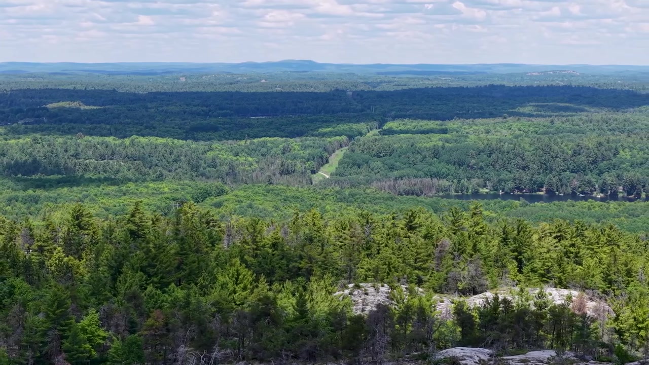 High aerial panorama sweeps across layered green forest and exposed granite ridges with a narrow clearing cutting through the northern wilderness
