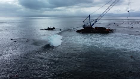 surfers ride the wild waves next to a crane shipwreck in bali, indonesia from an aerial drone shot