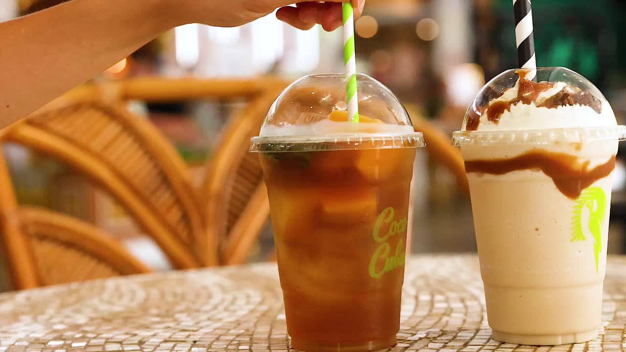 Two iced drinks on a table, one being stirred by hand. Bright, natural lighting enhances the vibrant setting