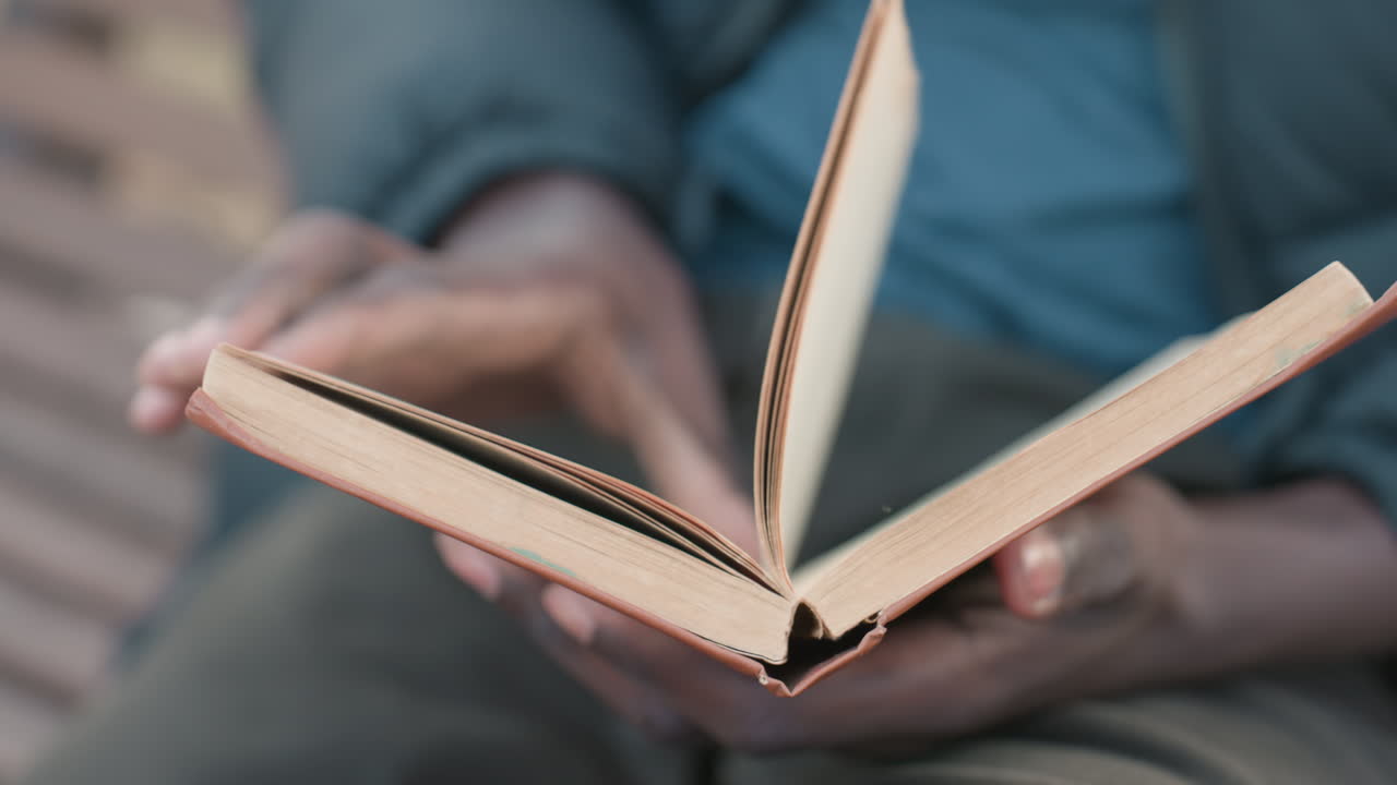 Close up of person reading novel outdoors, hands hold worn book on lap, pages mid turn, shallow depth of field with bench background
