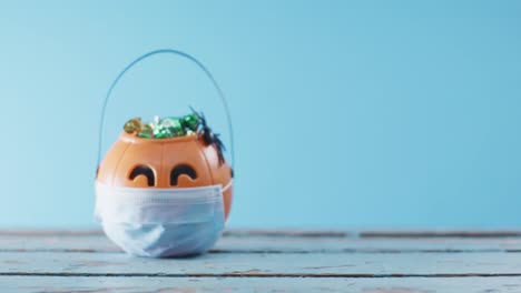 Face-mask-over-pumpkin-shaped-bucket-full-of-candies-on-wooden-surface-against-blue-background