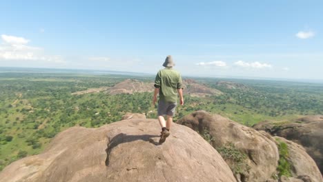 Aufnahme-In-Zeitlupe,-Die-Einen-Bärtigen-Ingwermann-Mit-Großem-Hut-Verfolgt,-Während-Er-Auf-Einen-Großen-Granitfelsen-Mit-Blick-Auf-Die-Ostafrikanische-Landschaft-Klettert