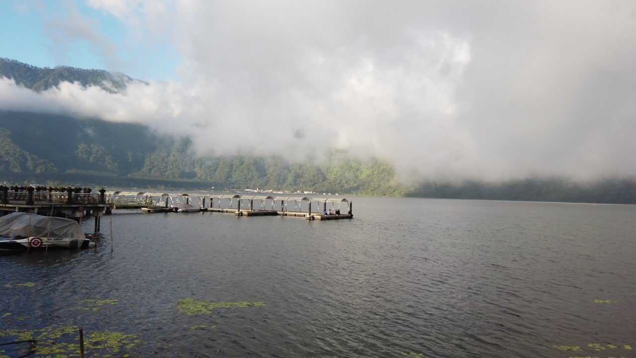 인도네시아 발리 에 있는 베라탄 호수 의 파노라마 뷰. 가 ⁇ 운 바람 이 불고, 안개 가 사라지고, 태양 이 배경 에 있는 산 들 을  ⁇ 고 들어가려고 한다.