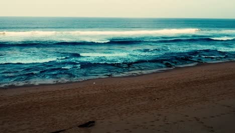 Luftaufnahmen,-Die-Mit-Einer-Drohne-Gefilmt-Wurden,-Mit-Blick-Auf-Den-Strand-Und-Das-Meer-Aus-Tosenden-Wellen-Mit-Einem-Tiefblauen-Ozean-Auf-Der-Klippe-In-Der-Südlichen-Hemisphäre-Afrikas