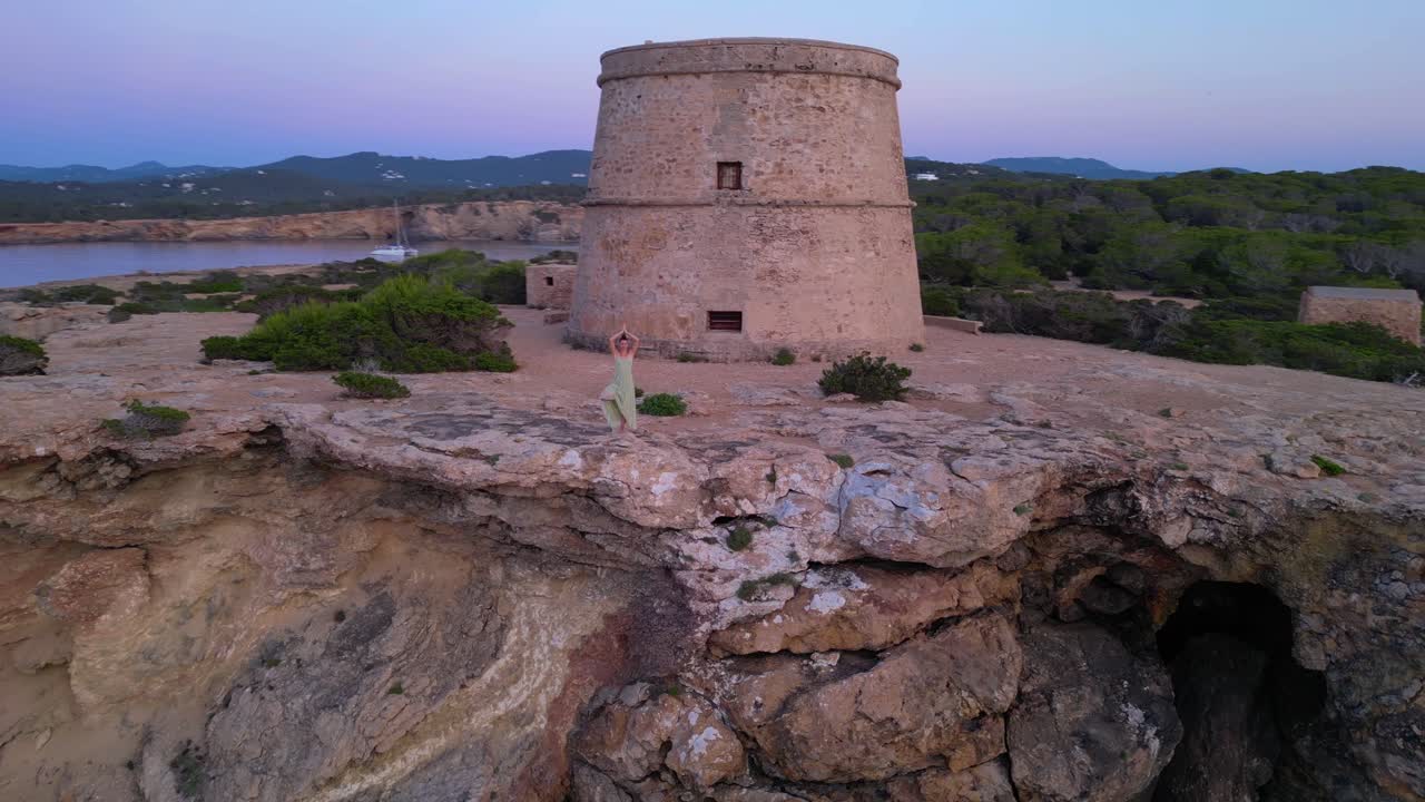 Barefoot hippie girl practicing yoga at sunset near Torre des Carregador, Ibiza, Spain. Beautiful aerial view flight wide orbit overview drone
