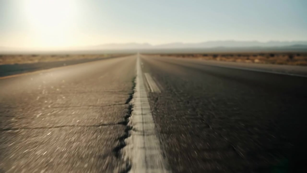 Low-angle roadside perspective of a straight two-lane highway cutting through flat arid terrain toward a distant mountain range. Textured asphalt with cracks