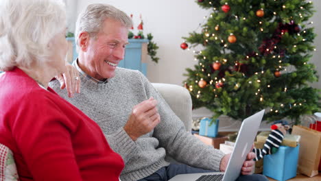 Nahaufnahme-Eines-älteren-Paares,-Das-Auf-Dem-Sofa-Sitzt-Und-Mit-Einem-Laptop-Zu-Hause-Waren-Oder-Dienstleistungen-Kauft,-Mit-Einem-Weihnachtsbaum-Im-Hintergrund