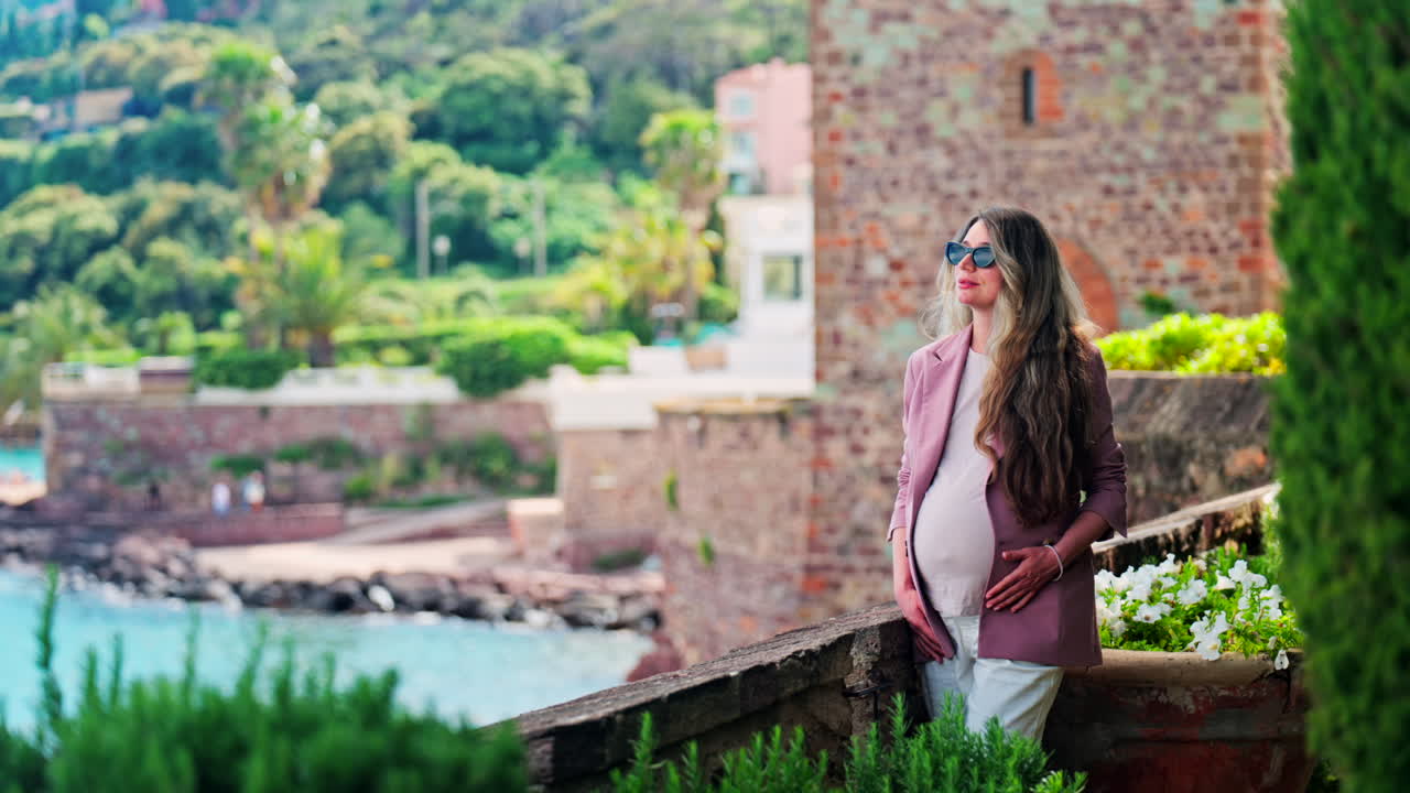 Pregnant woman with long hair, wearing sunglasses standing with the Chateau de la Napoule Castle in Mandelieu-La Napoule, France in the background