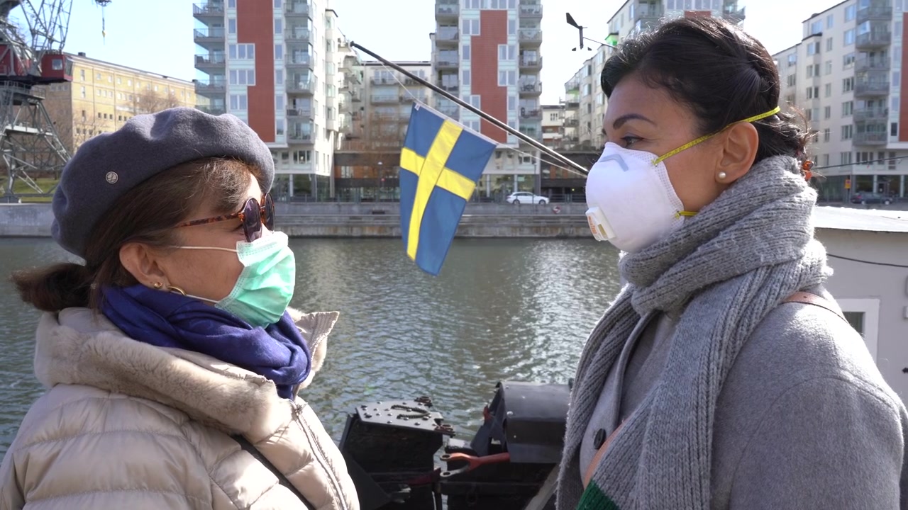 Aasialaisen äidin ja tyttären muotokuva, he katsovat toisiaan ja käyttävät suojaavaa kasvomaskia koronavirusepidemiaa vastaan. Taustalla sininen meri ja Ruotsin lippu. Virussuojaus, Ruotsi.